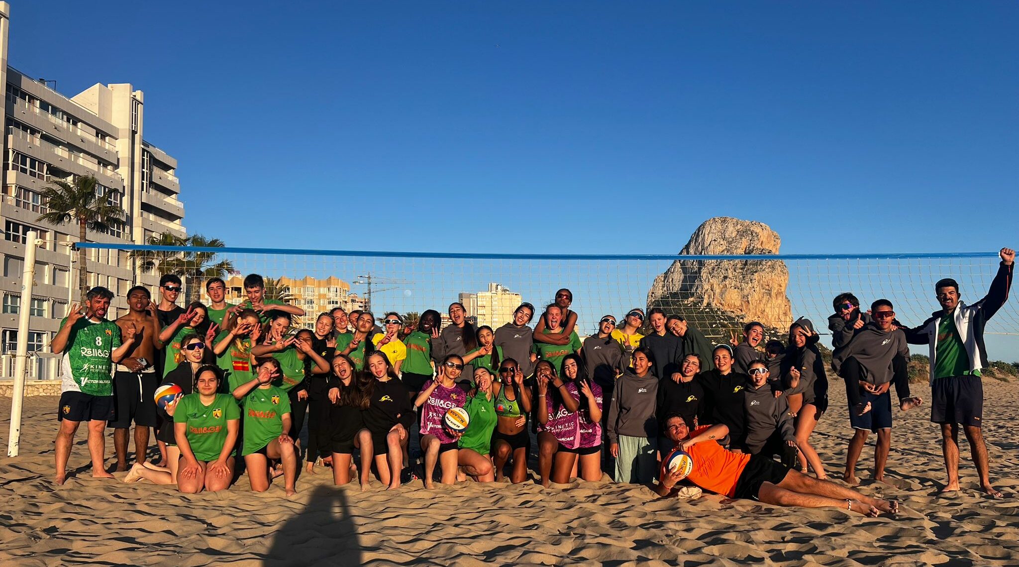 Foto de grupo de jugadores, jugadoras y entrenadores del Club Voleibol Ifach y Club Voley Playa Las Rozas durante el campus en la playa con el Peñón de Ifach al fondo